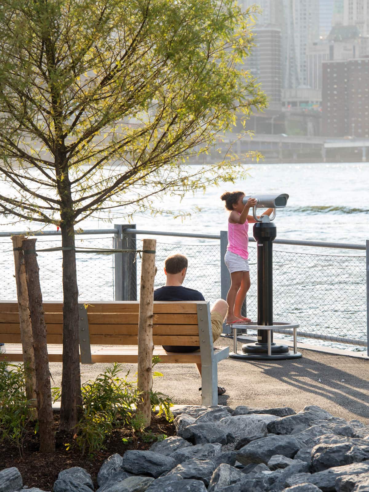 Man sitting on bench and girl looking through binoculars by the water on a sunny day.