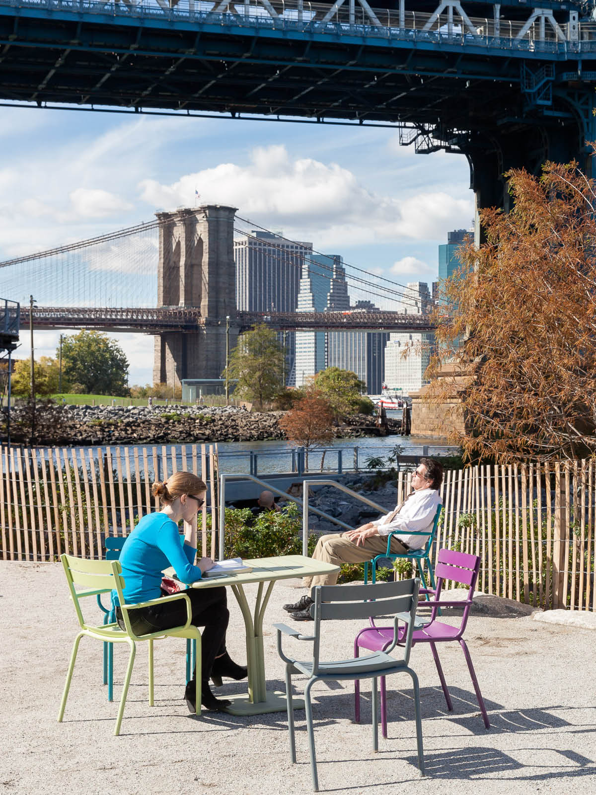 People sitting at tables on Pearl Street Terrace on a sunny day. Manhattan Bridge is seen overhead and Brooklyn Bridge is seen in the distance.