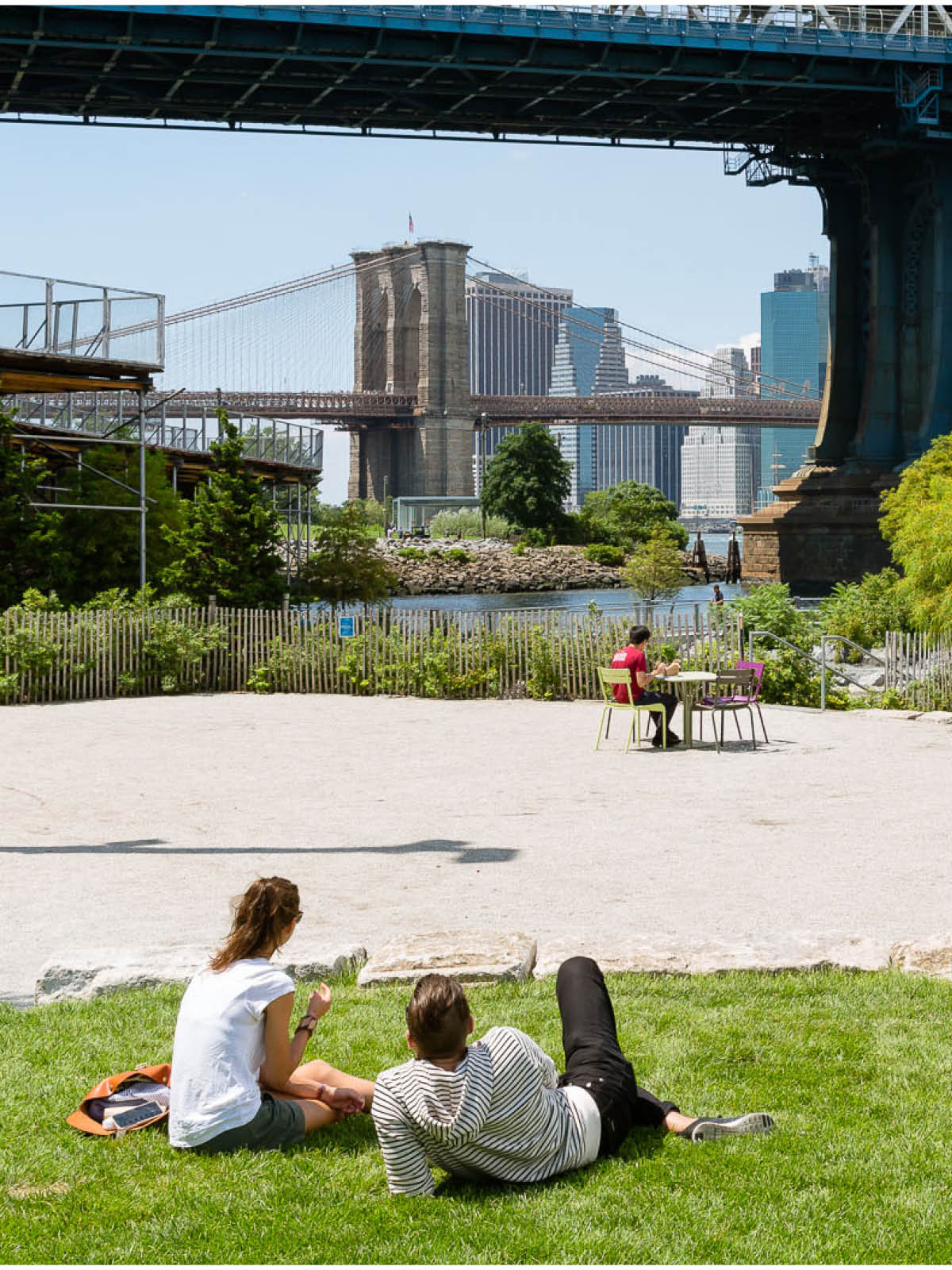 Couple sitting on grass next to Pearl Street Terrace on a sunny day. Manhattan Bridge is seen overhead and Brooklyn Bridge in the distance.