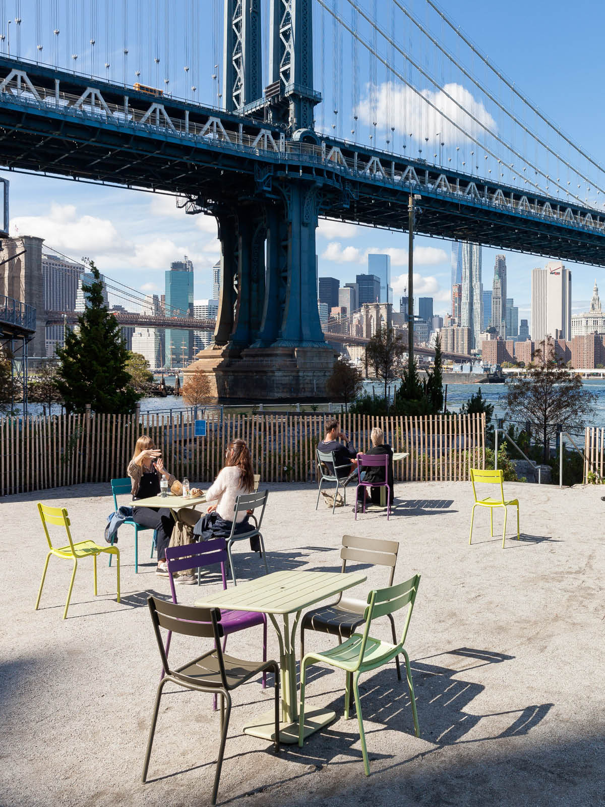 Groups sitting at tables on a terrace on a sunny day. The Manhattan Bridge is seen in the background.