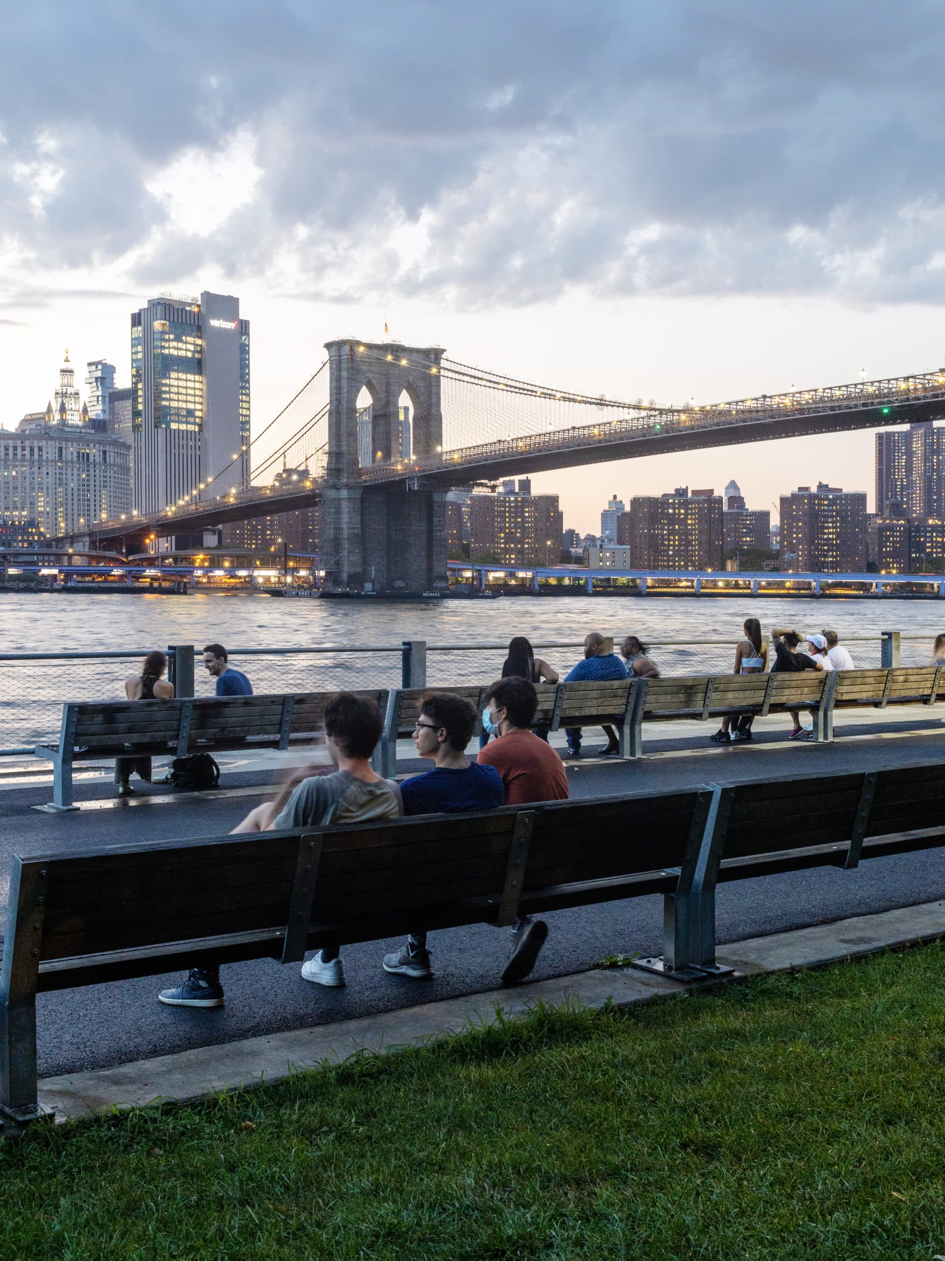 Peope sitting on benches on the Pier 1 Promenade looking out at the Brooklyn Bridge at sunset.