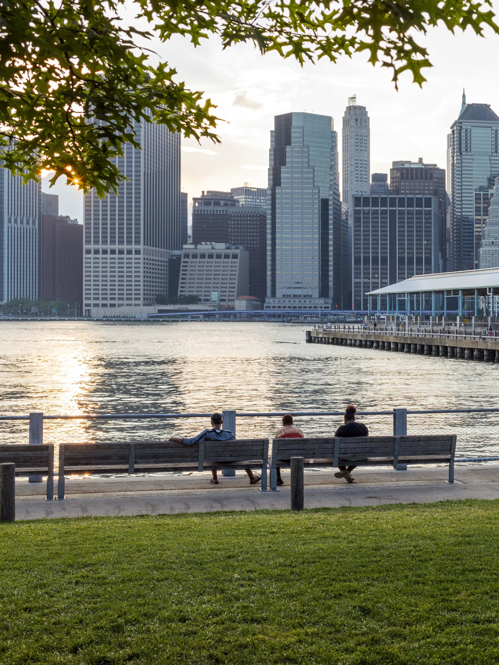 People sitting on benches by the water with a view of Pier 2 and lower Manhattan at sunset.