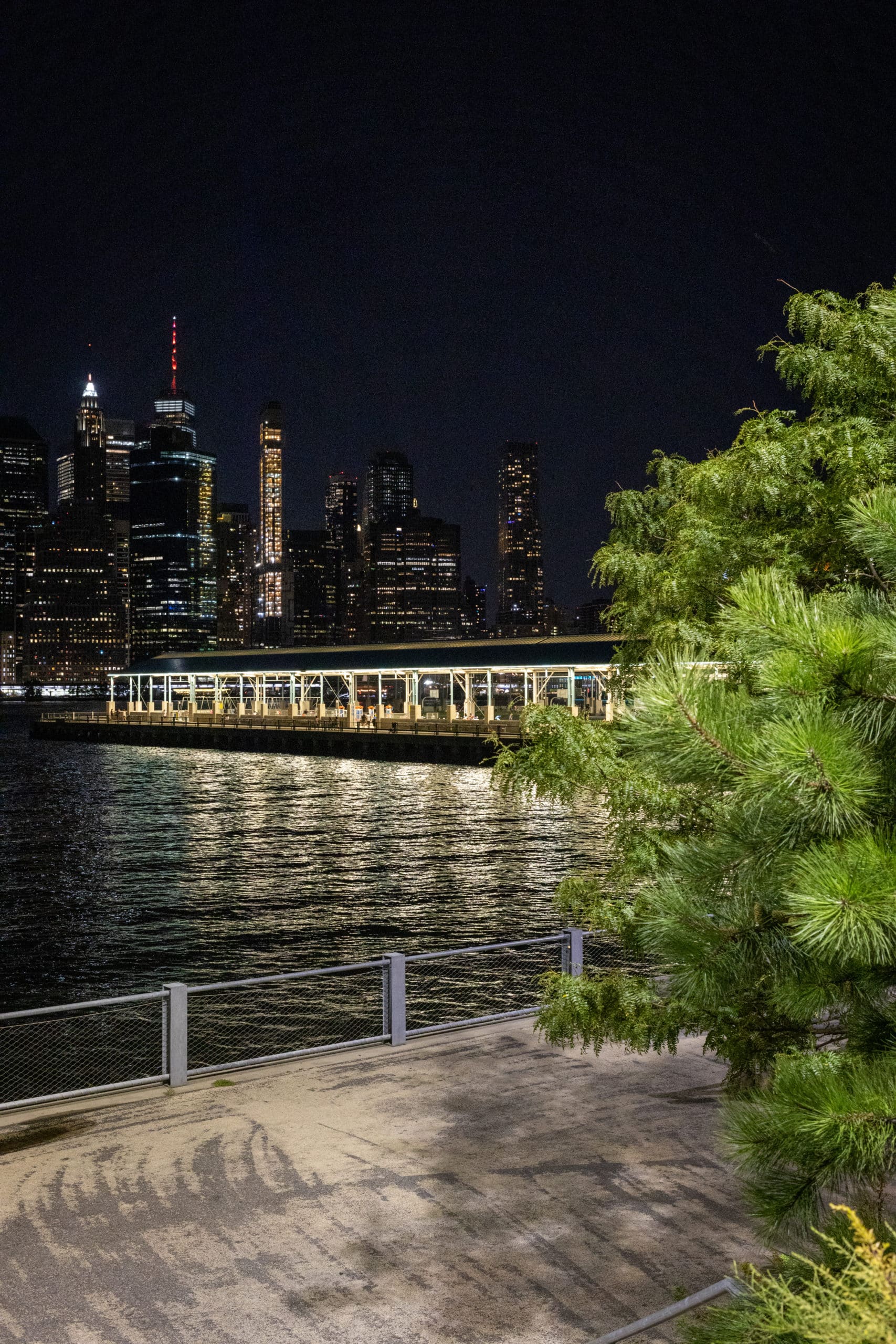 Pathway by the water on Pier 3 Greenway with a view of Pier 2 and lower Manhattan at night.