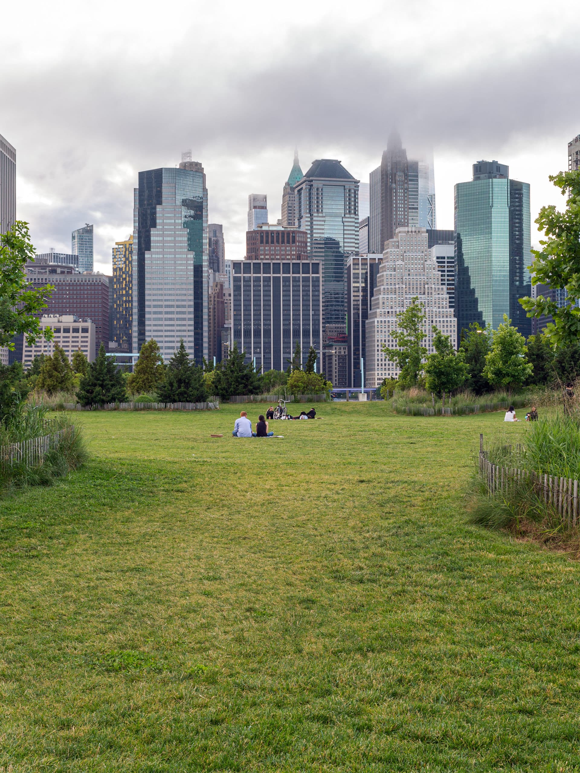 People sitting far off on the Pier 3 Lawn on a cloudy day. Lower Manhattan is seen in the background.