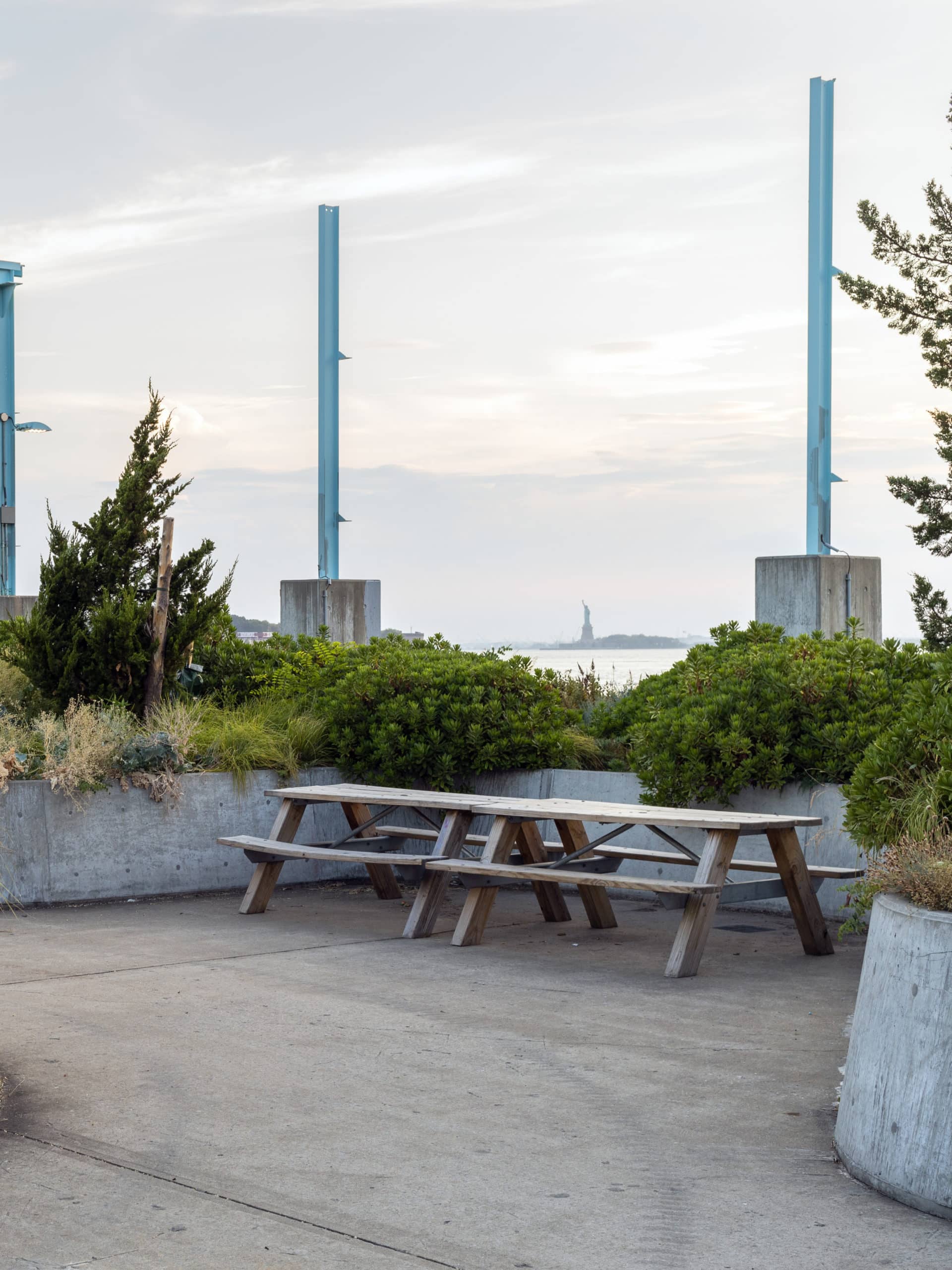 Picnic tables beside garden areas on Pier 3 at sunset. The Statue of Liberty is seen in the distance.