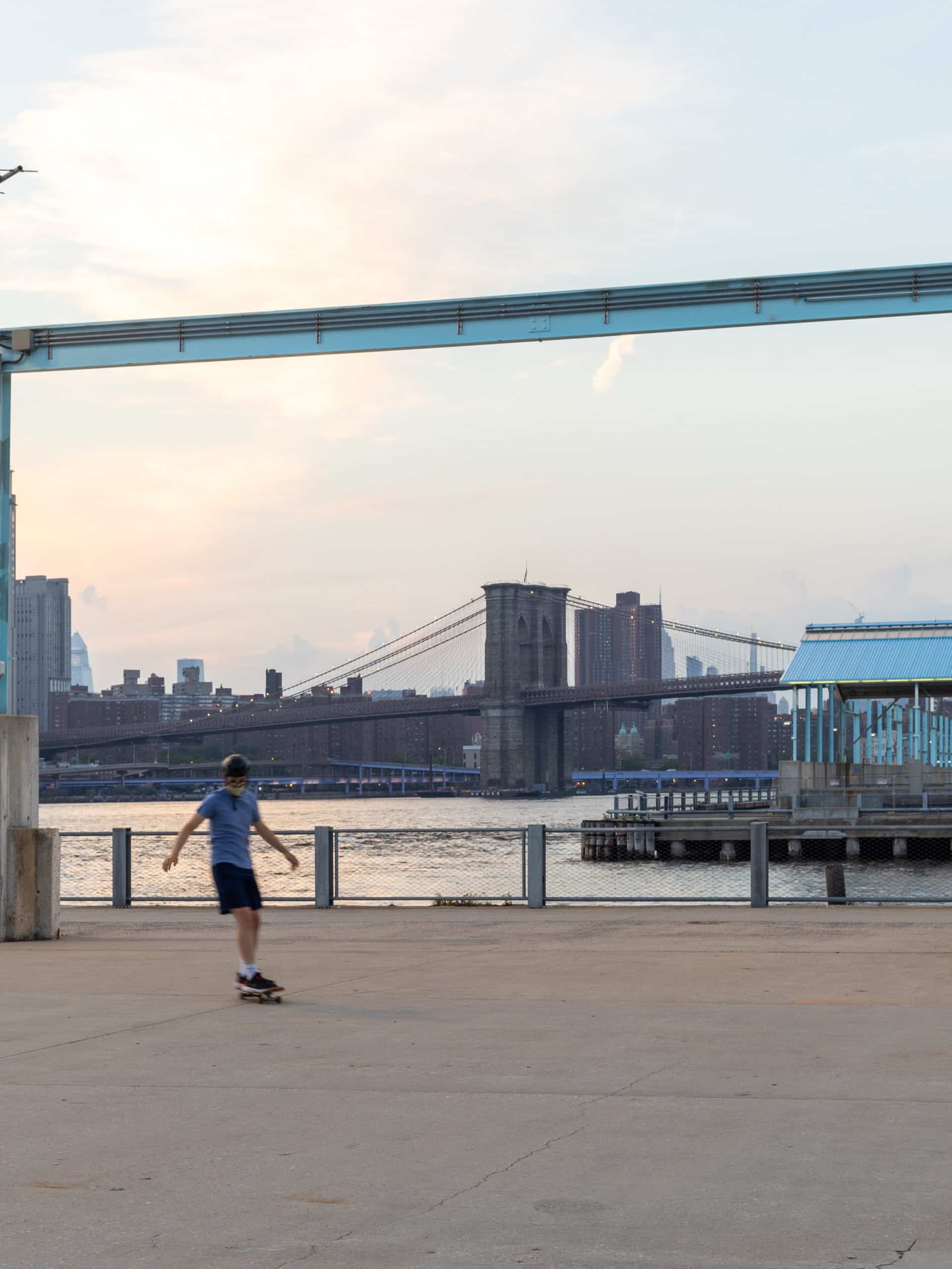 Boy skateboarding on Pier 3 Plaza with Pier 2 and Brooklyn Bridge in background at sunset.