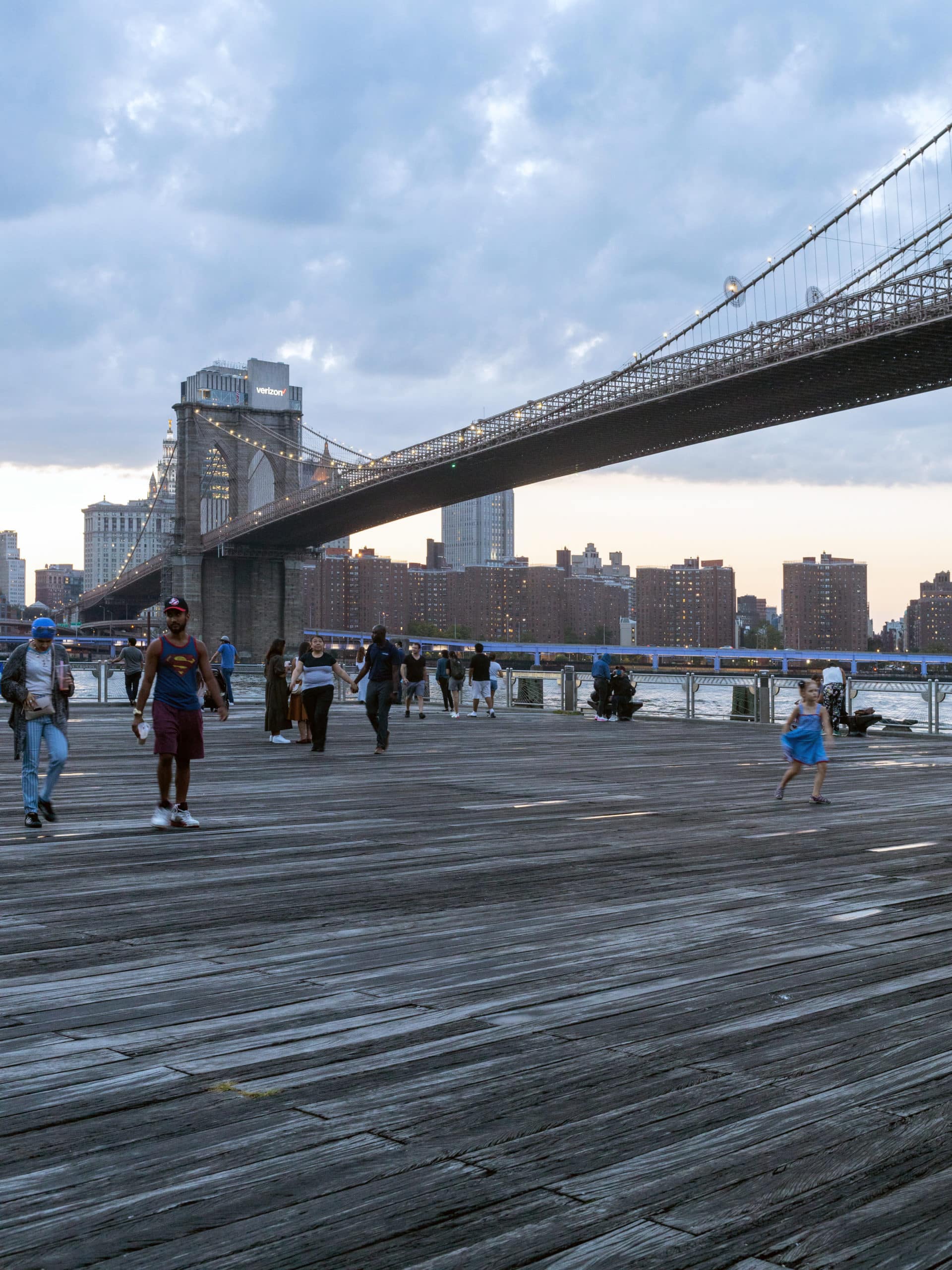 People on the Fulton Ferry Landing Boardwalk at sunset. View of the Brooklyn Bridge and the Lower East Side in the background.