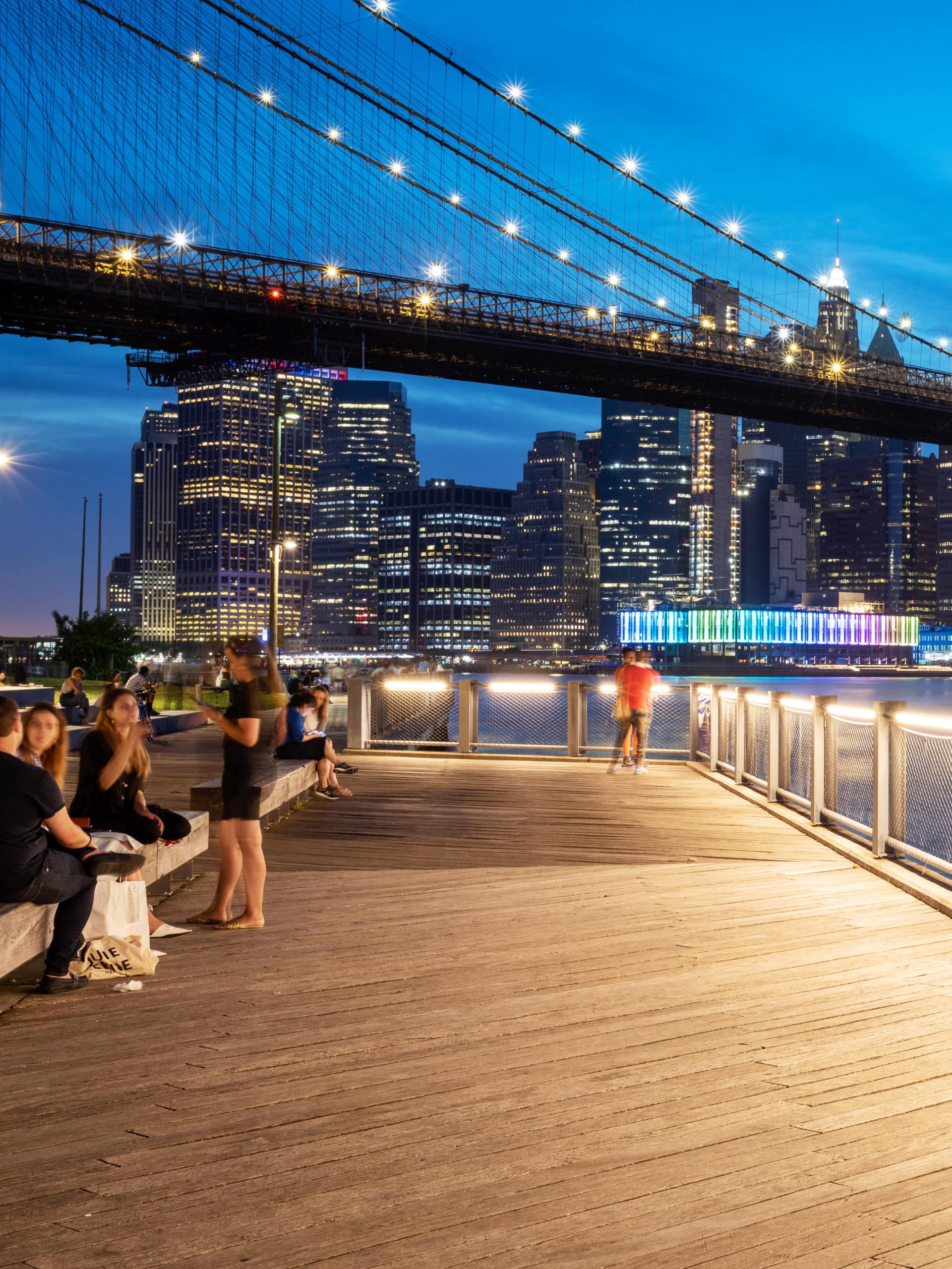 People sitting on the boardwalk at night with a view of the Brooklyn Bridge and Pier 17 in Manhattan.