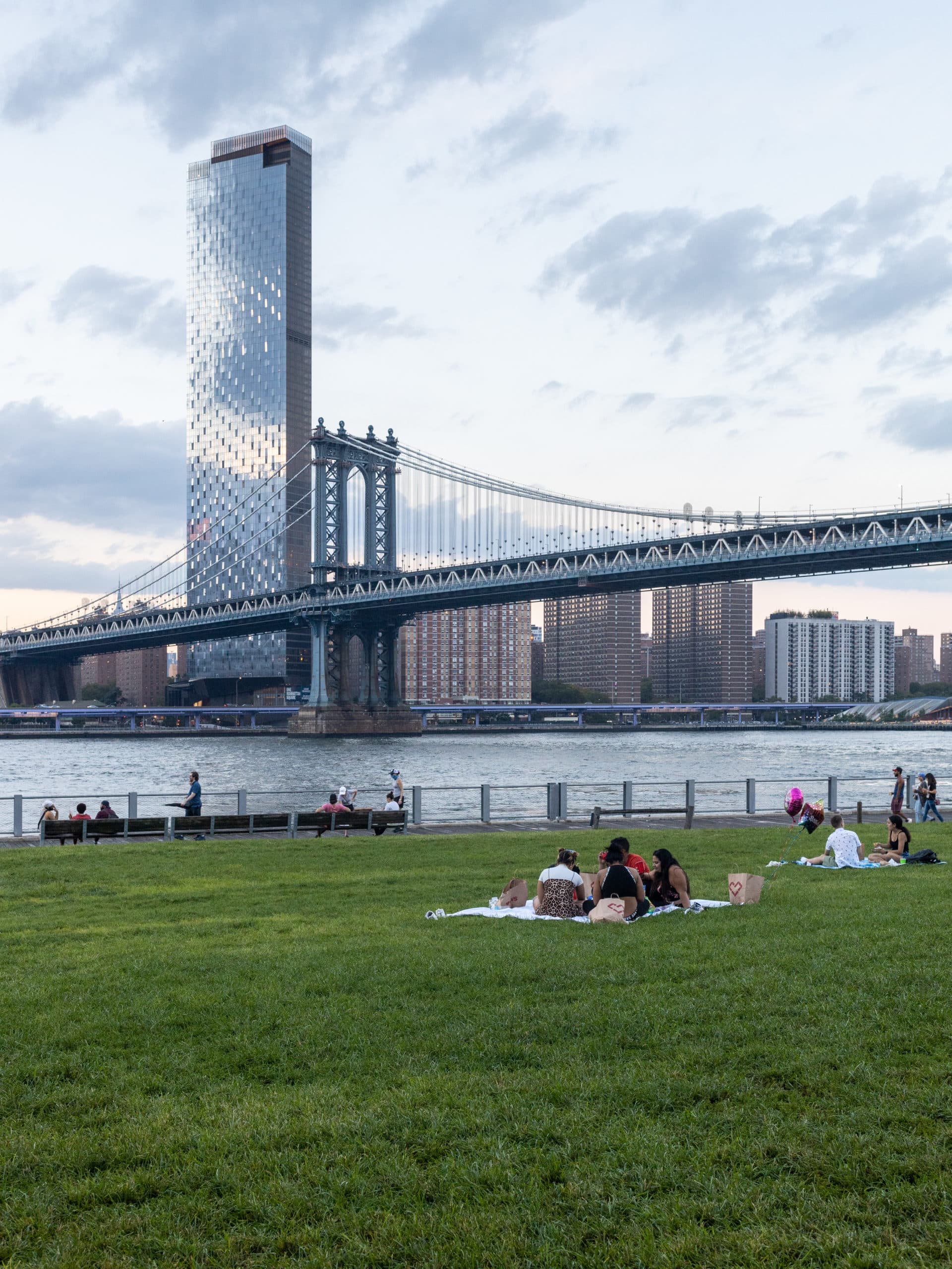 Group sitting on the Empire Fulton Ferry Lawn at dusk with the East River and Manhattan Bridge in background.