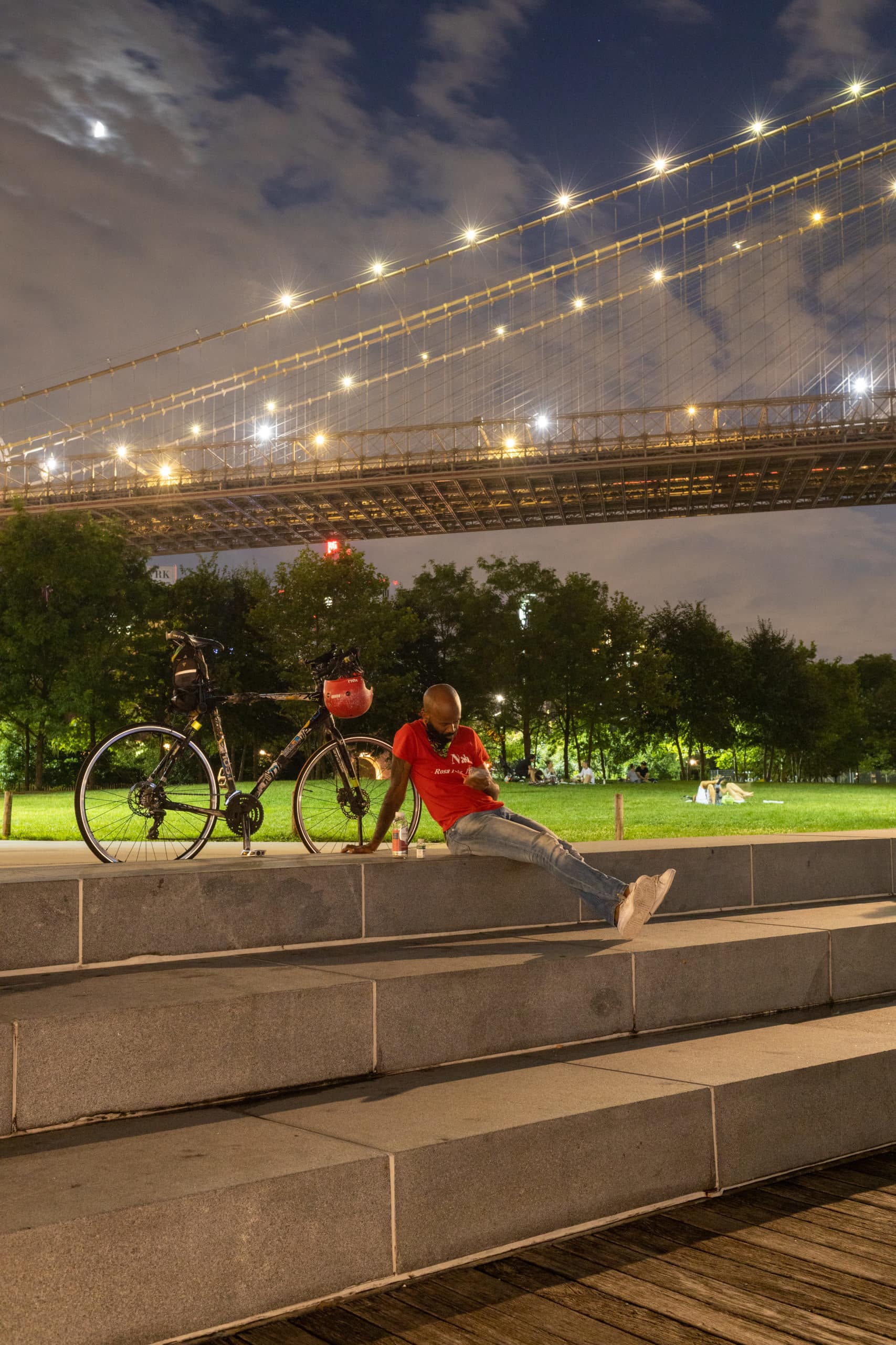 Man with a bike sitting on steps by Empire Fulton Ferry Lawn at night with Brooklyn Bridge overhead.