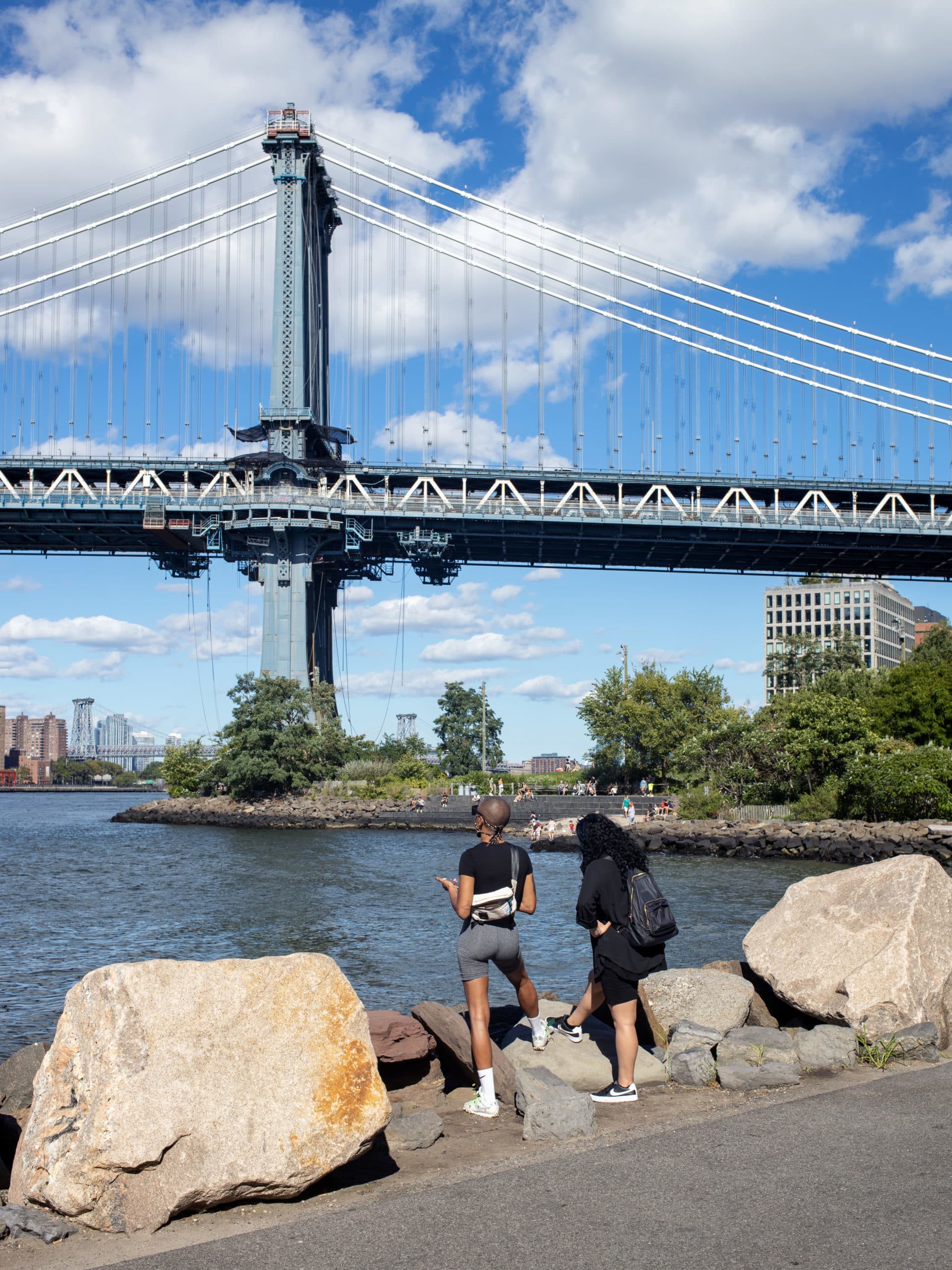People on the pathway by the water looking up at the Manhattan Bridge on a sunny day.