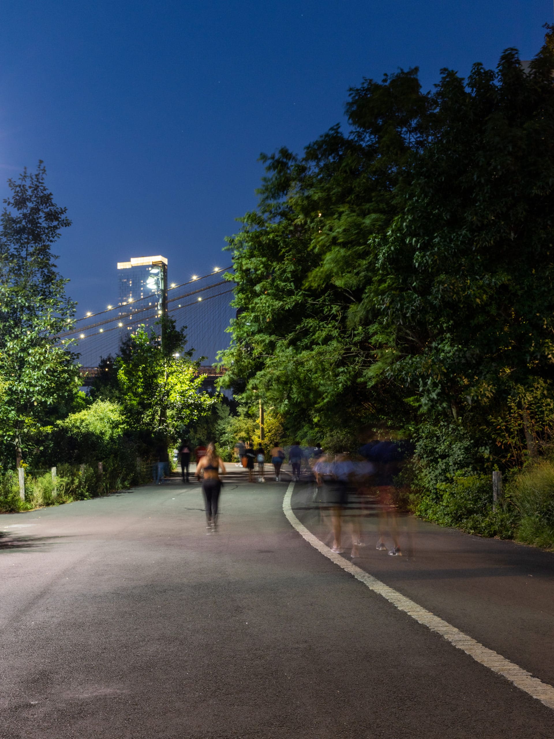 Joggers running on the Greenway at night with the Brooklyn Bridge and One Manhattan Square in the distance.