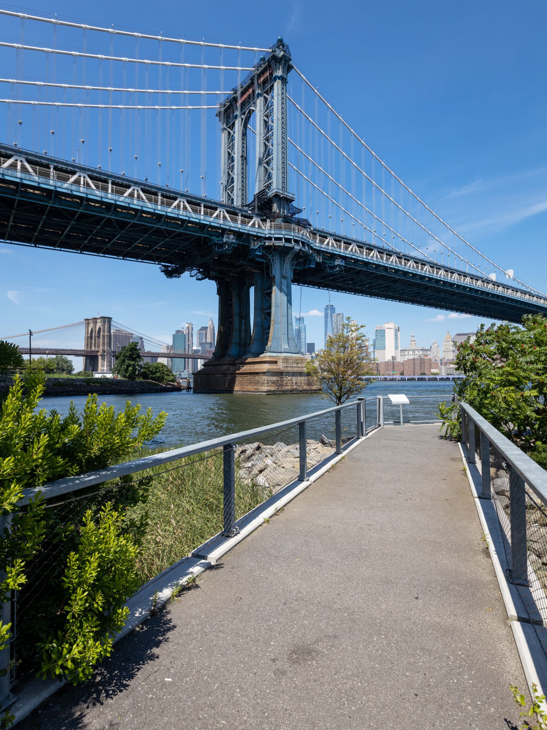 Pedestrian Bridge by the water on a sunny day. The Manhattan and Brooklyn Bridges are seen in the distance.