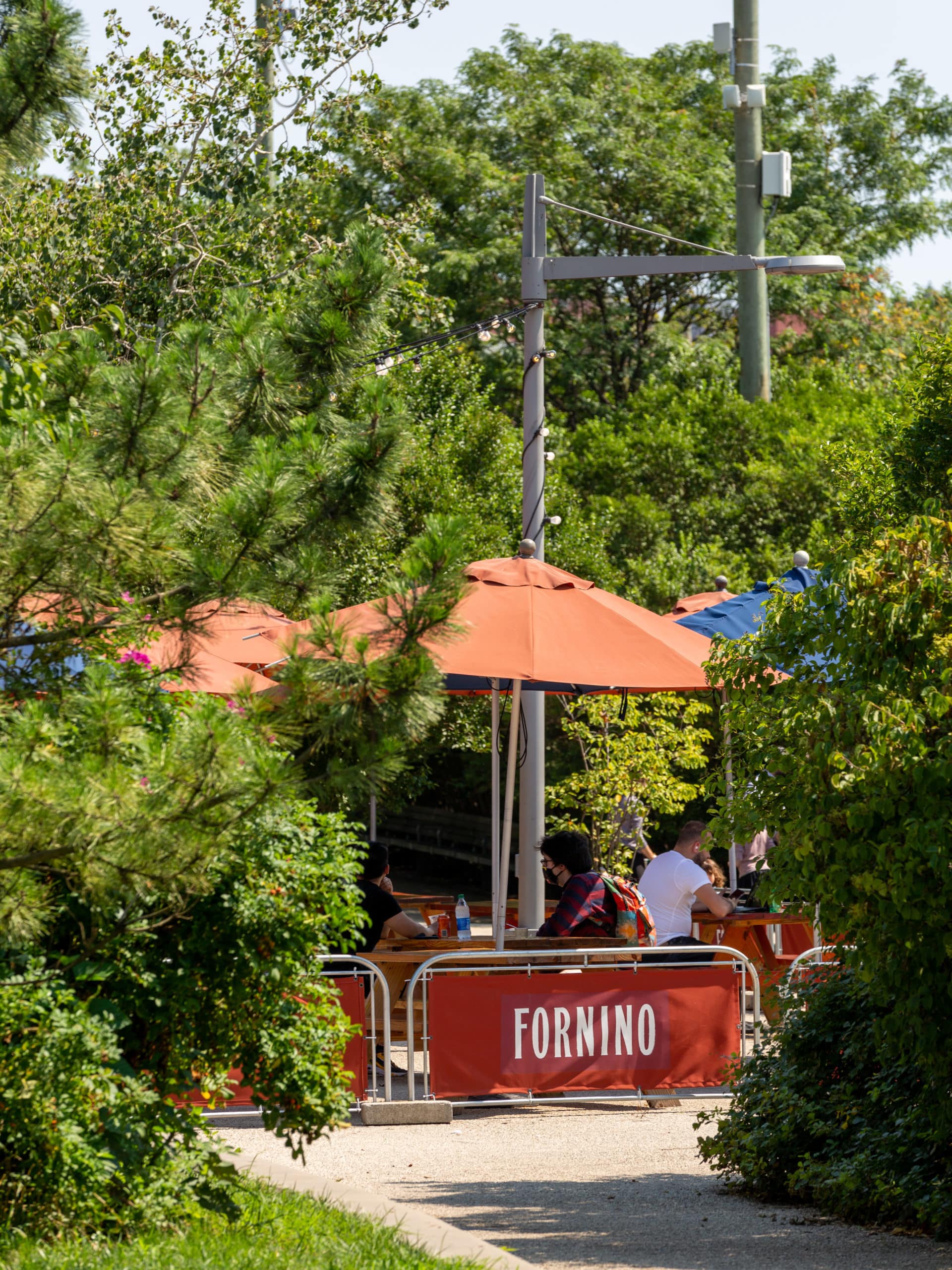 View from tree-lined pathway of people sitting under umbrellas at Fornino on a sunny day.