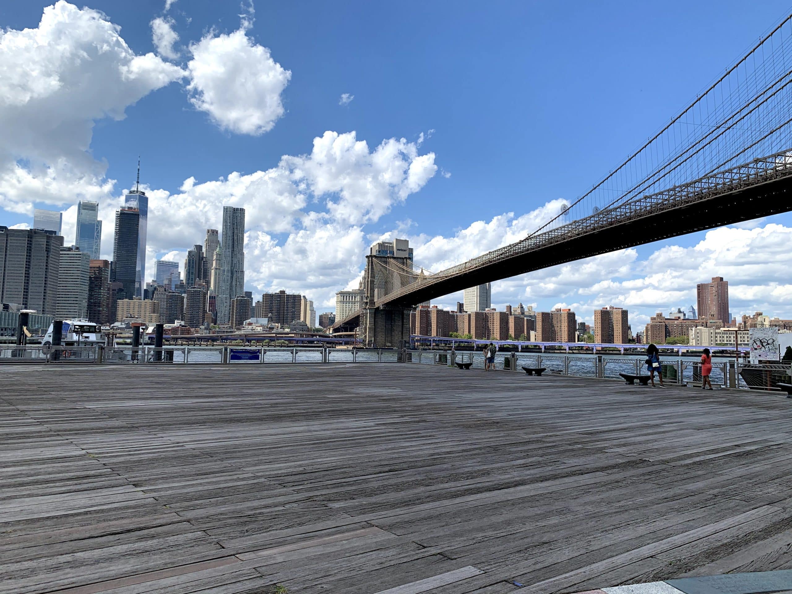 Fulton Ferry Landing boardwalk with views of the Brooklyn Bridge and lower Manhattan on a cloudy day.