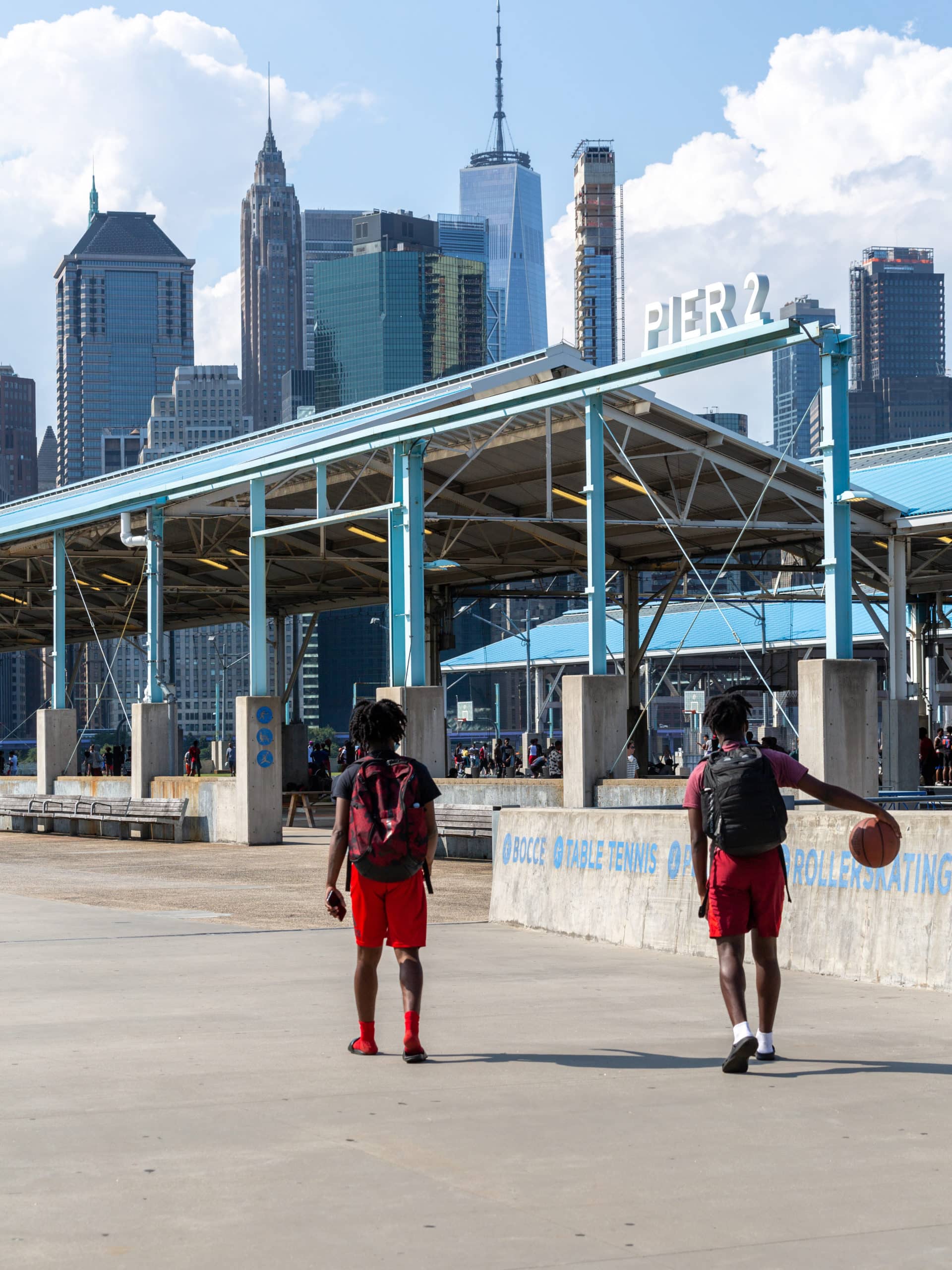 A man dribbles a basketball as he and another man walk towards Pier 2 on a sunny day.