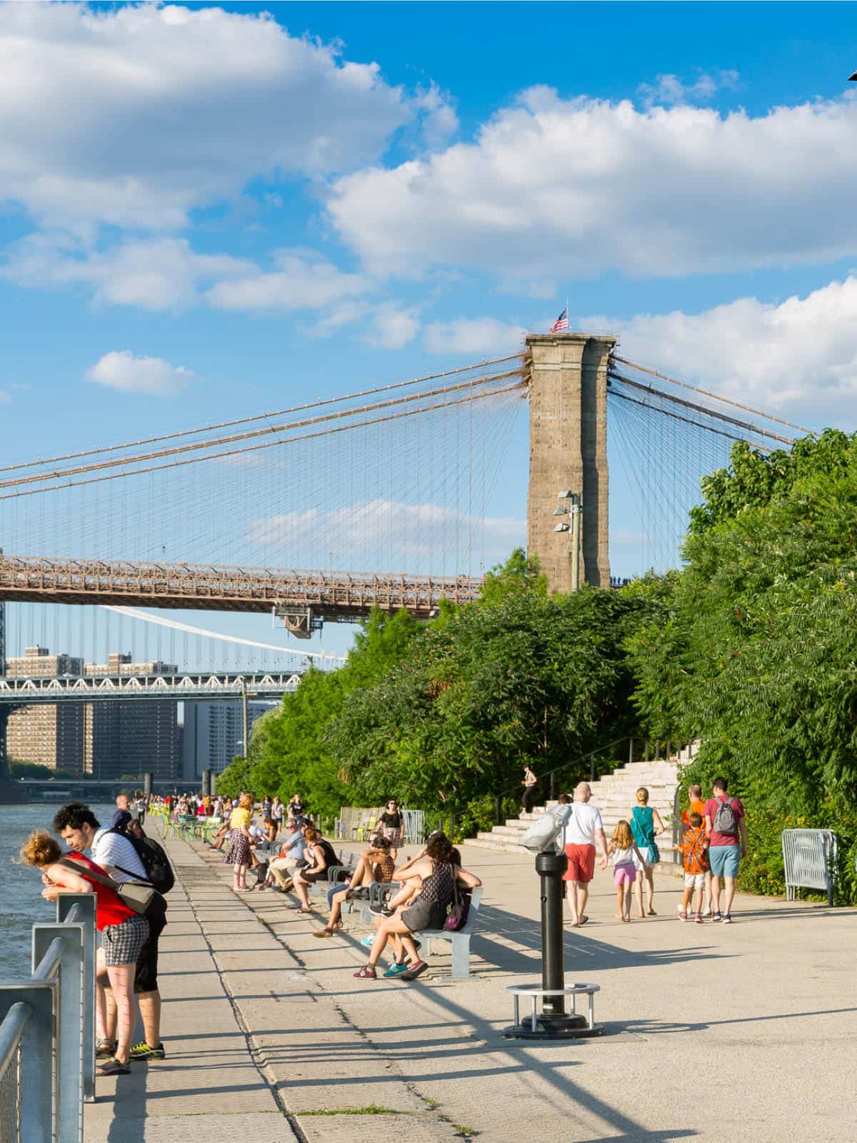 People sitting on promenade on a sunny day. The Brooklyn Bridge is seen in the background.