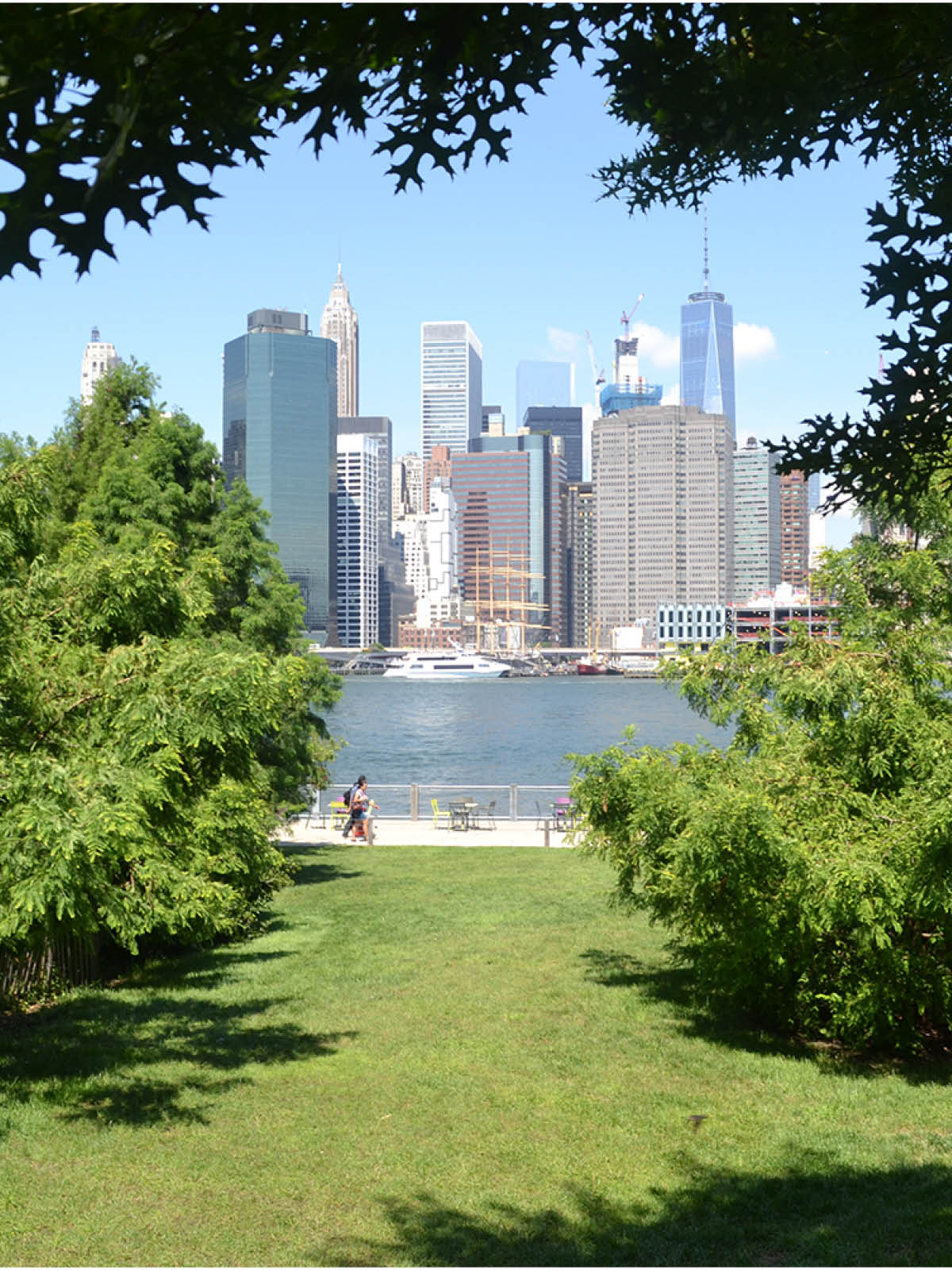 Lawn surrounded by trees with views of water and lower Manhattan on a sunny day.