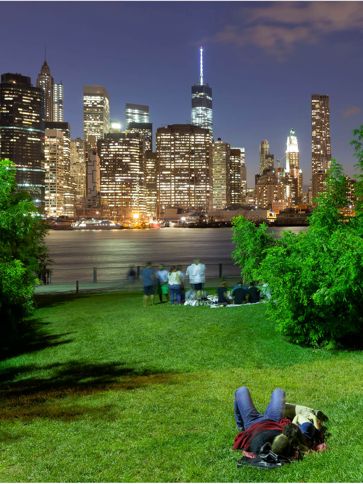 Couple lying on grass at night on the Pier 1 Lawn. Lower Manhattan is seen in the background.