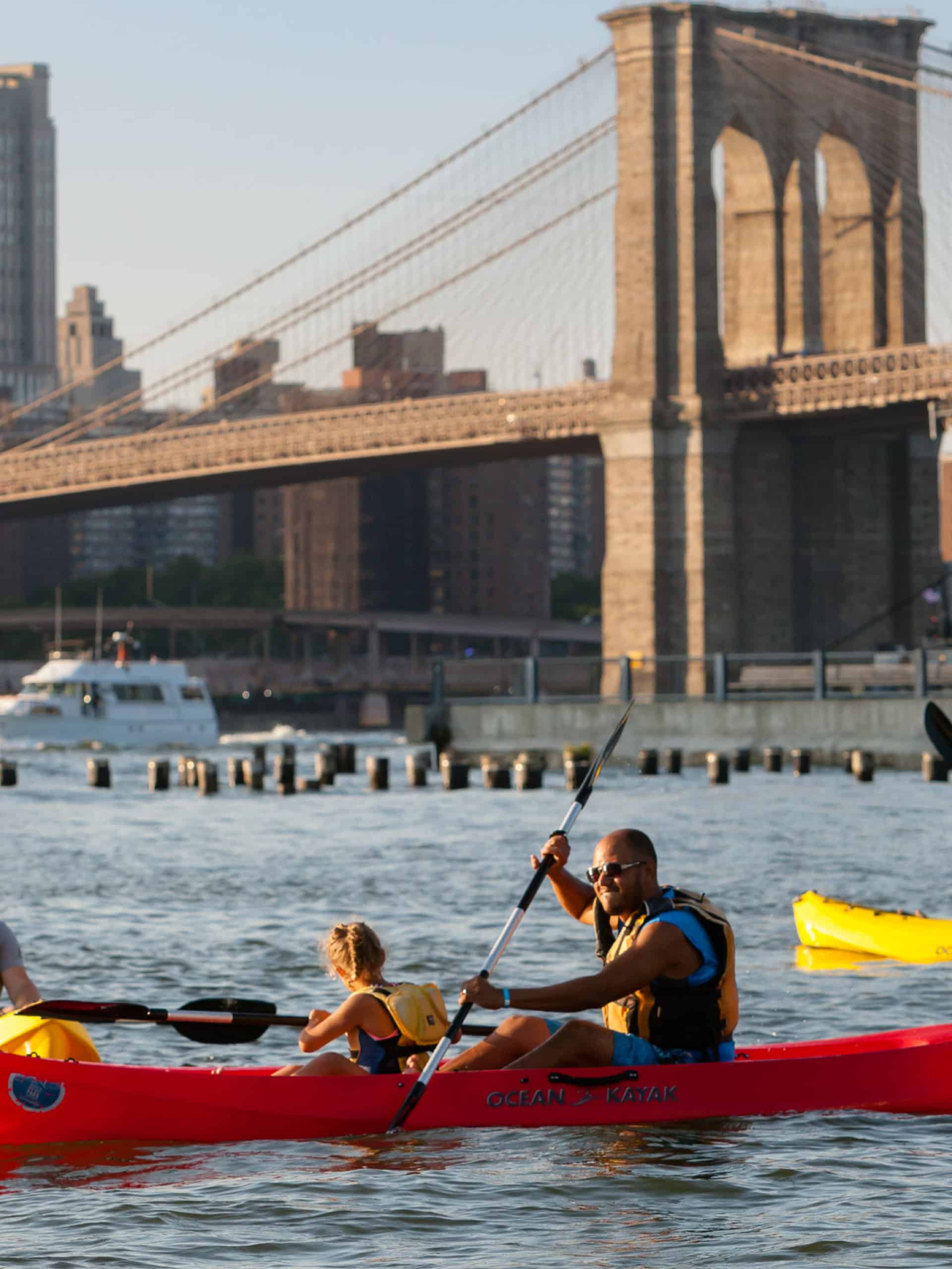 Family kayaking on a sunny day with the Brooklyn Bridge in the background.