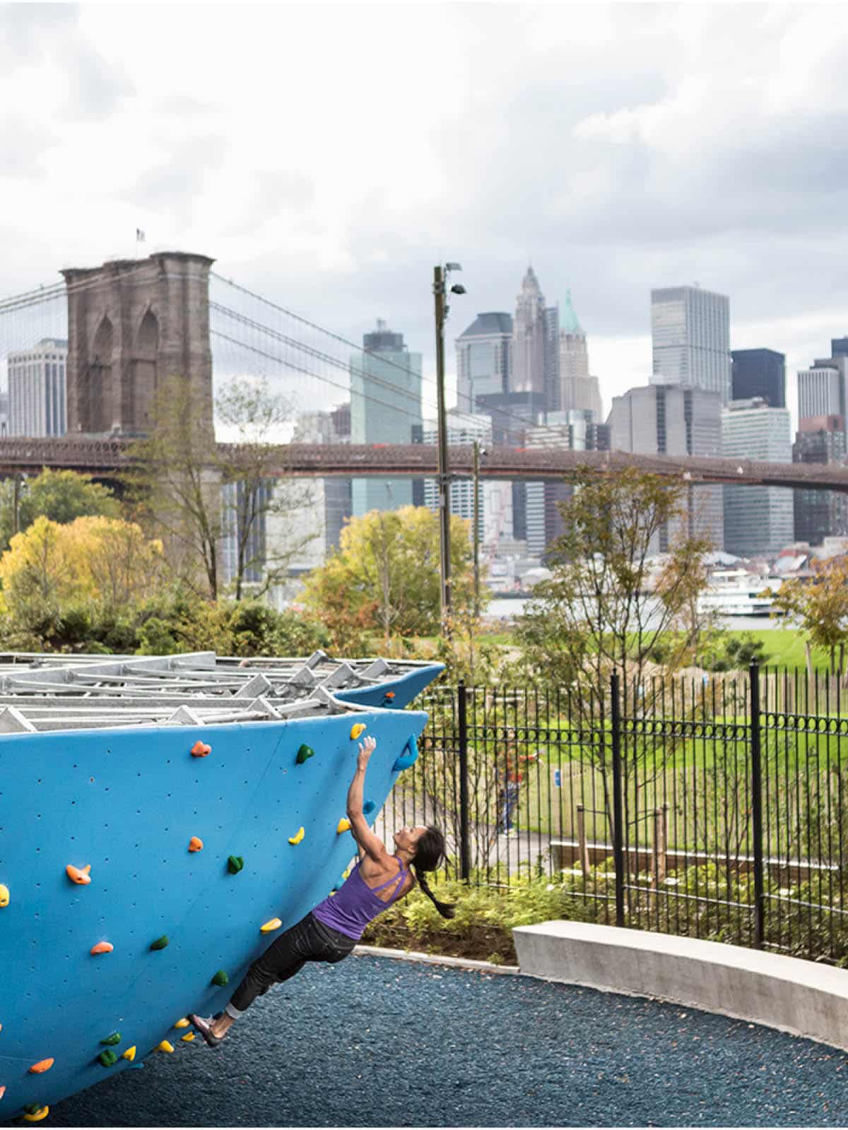 Woman climbing a bouldering wall on a cloudy day with the Brooklyn Bridge in the background.