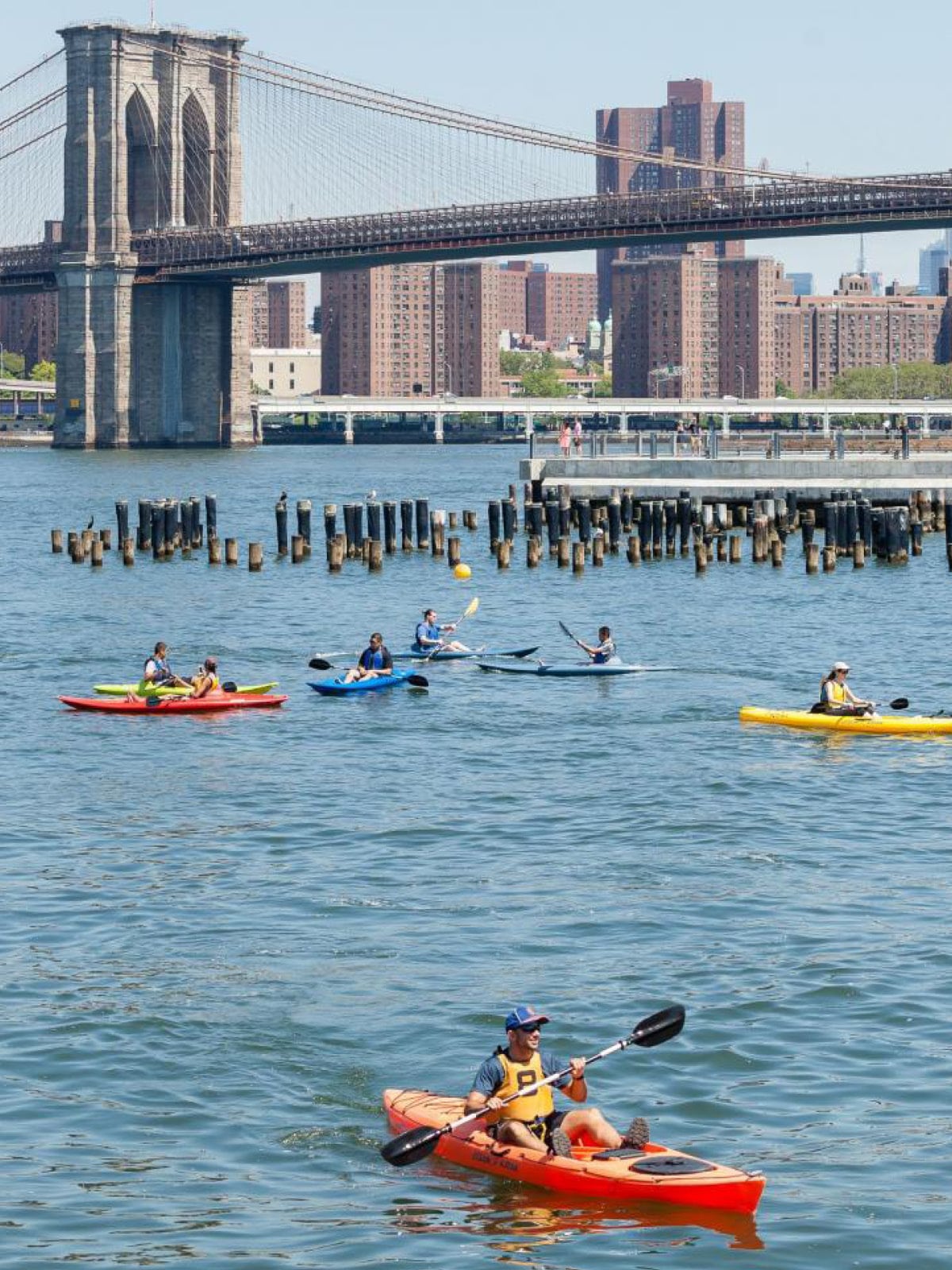Man kayaking with other kayakers in behind on a sunny day. The Brooklyn Bridge is seen in the distance.