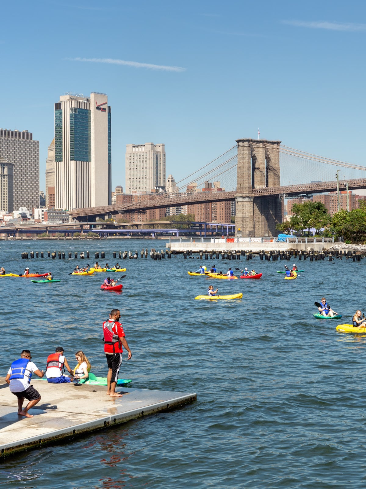 People standing on the dock looking at kayakers in the water on a sunny day. The Brooklyn Bridge is seen in the background.