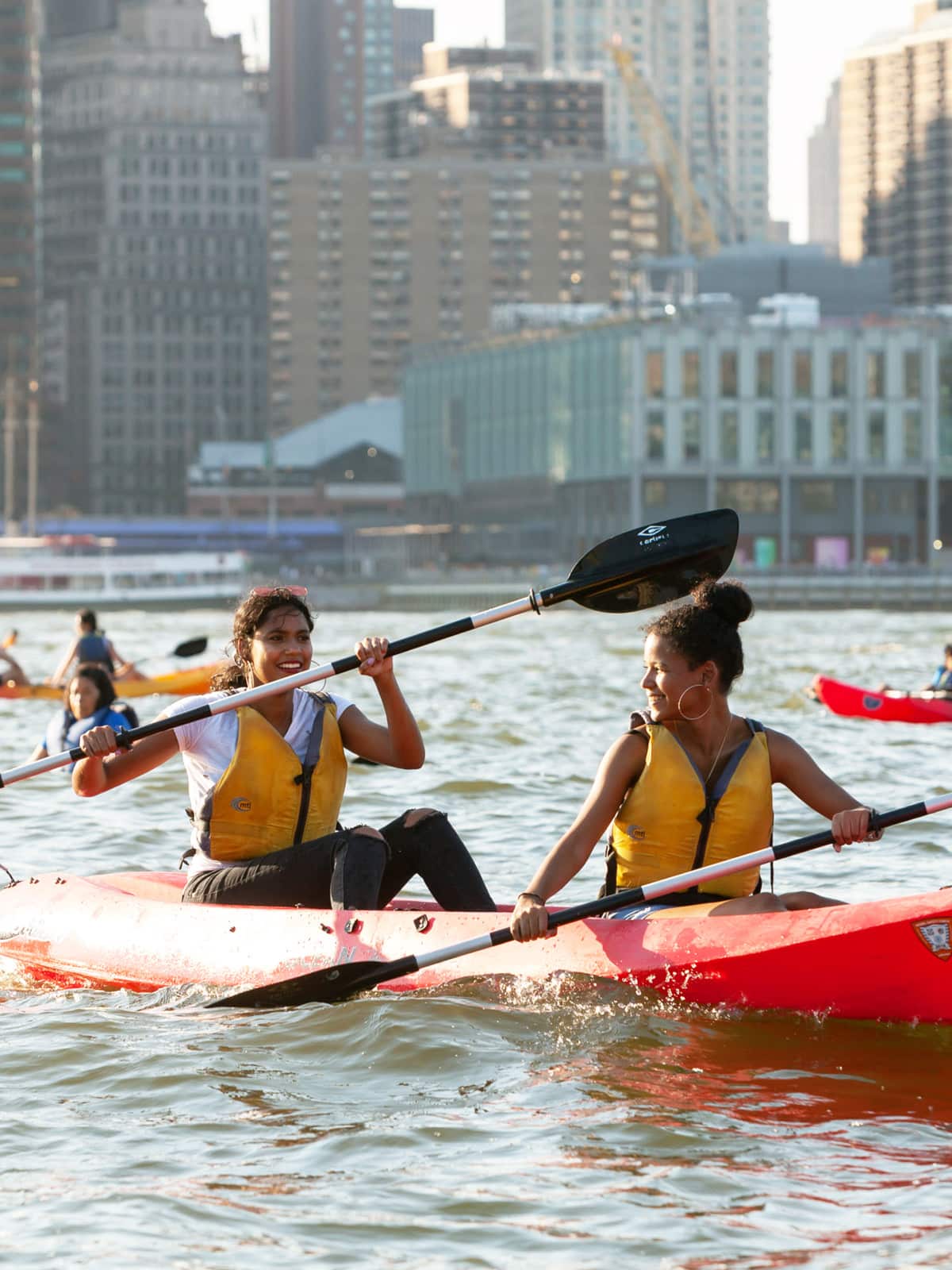 Close up of two women in a kayak on a sunny day. Pier 17 is seen in the background.