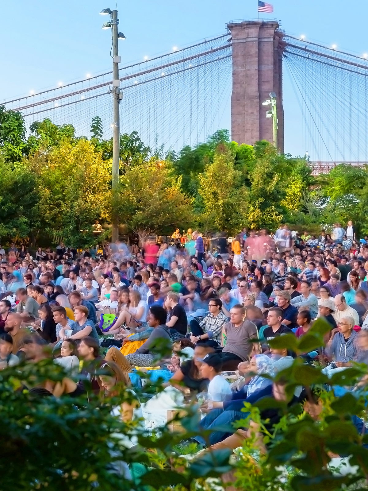 Crowd sitting on the lawn watching a movie at night. The Brooklyn Bridge is seen in the distance.