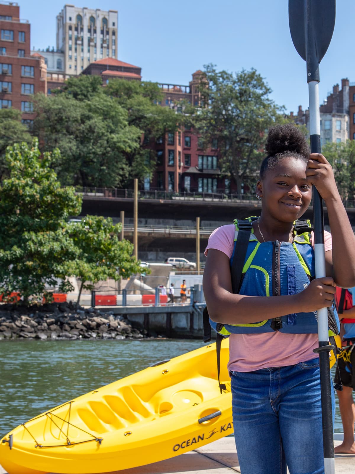 Close up of girl with kayak paddle with a kayak in the background on a sunny day. Brooklyn Heights is seen in the distance.