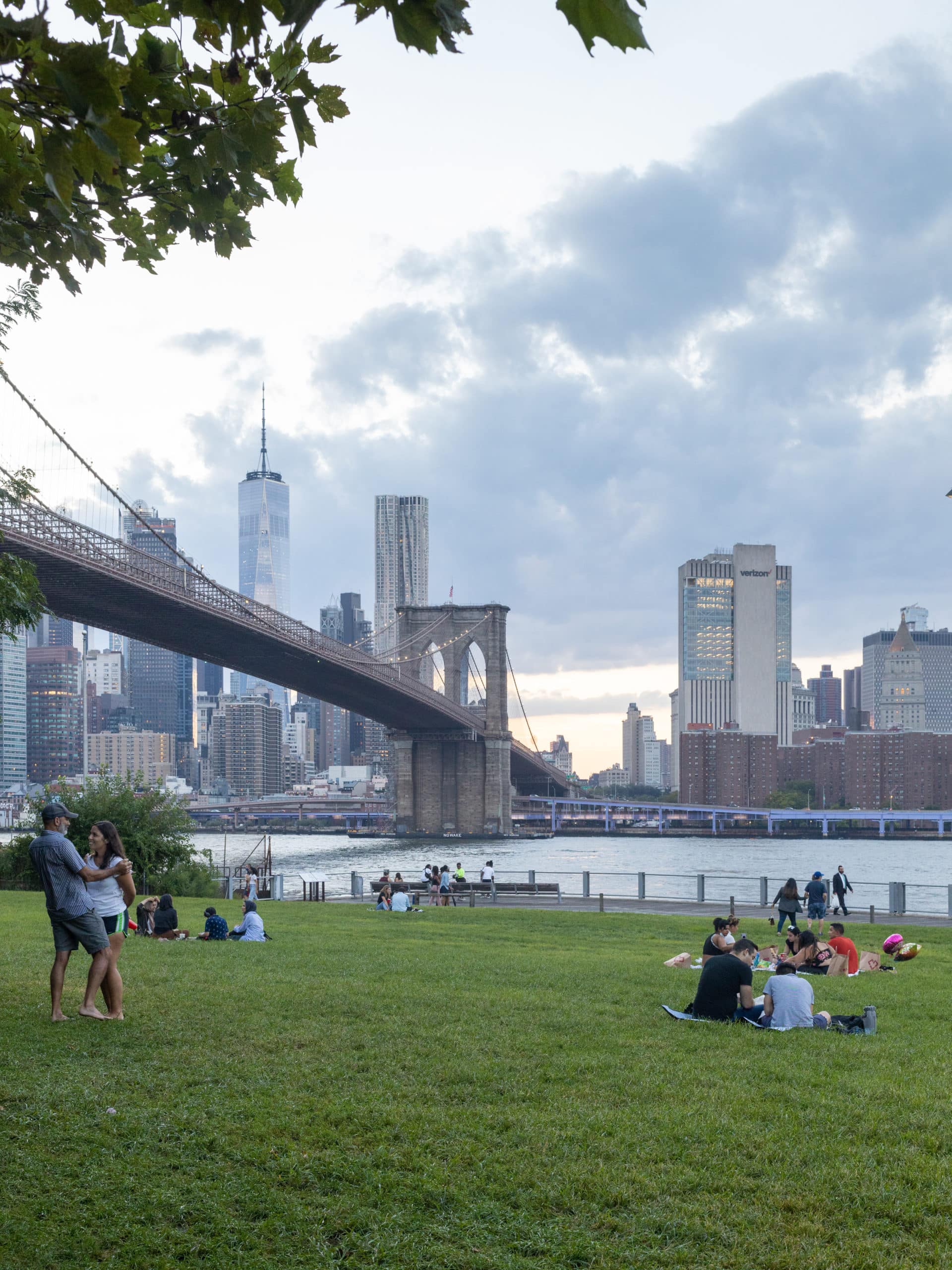 People sitting on Empire Fulton Ferry Lawn at dusk with view of the Brooklyn Bridge and lower Manhattan.