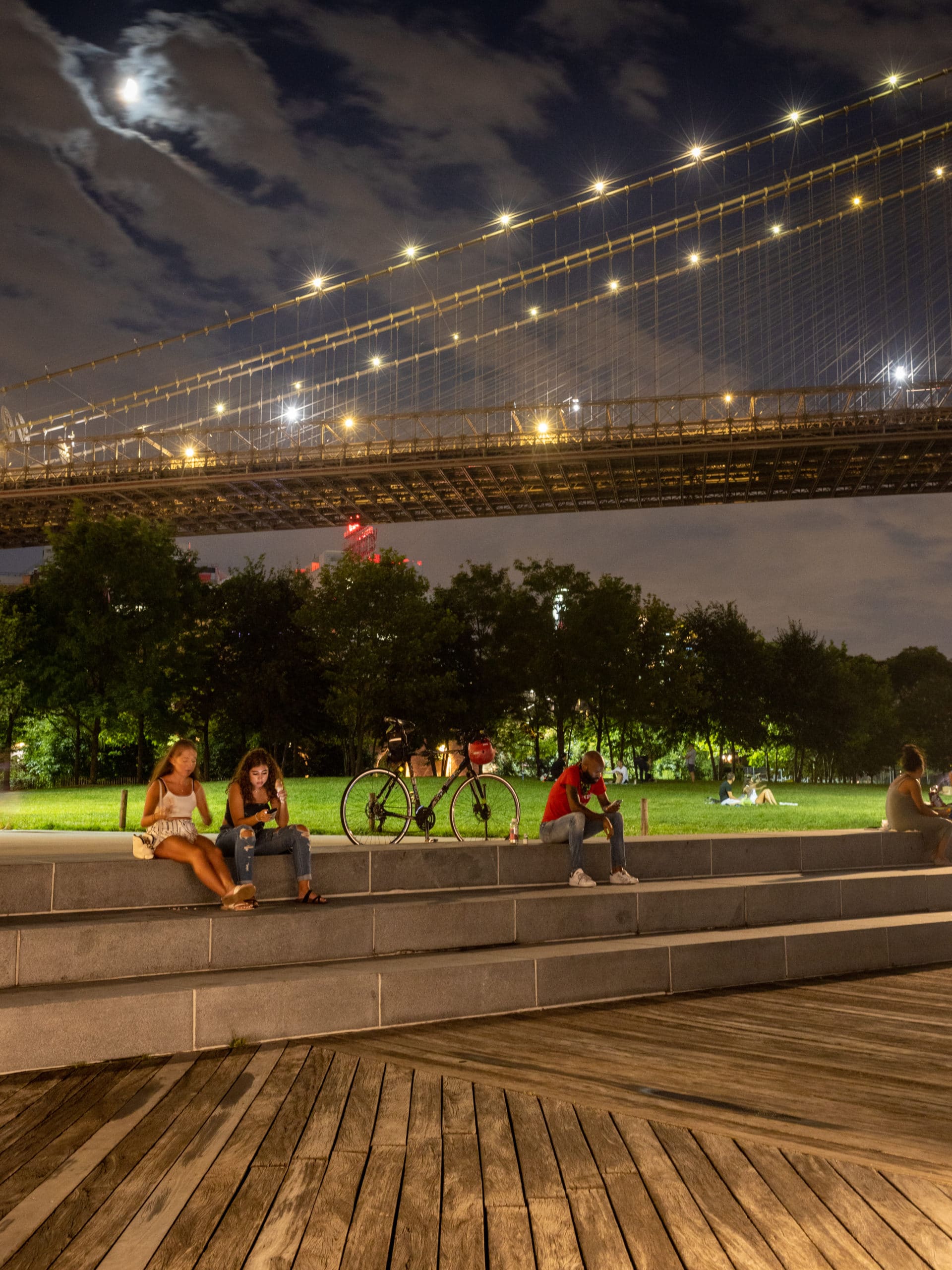 People sitting on steps by the Empire Fulton Ferry Lawn at night with the Brooklyn Bridge overhead.