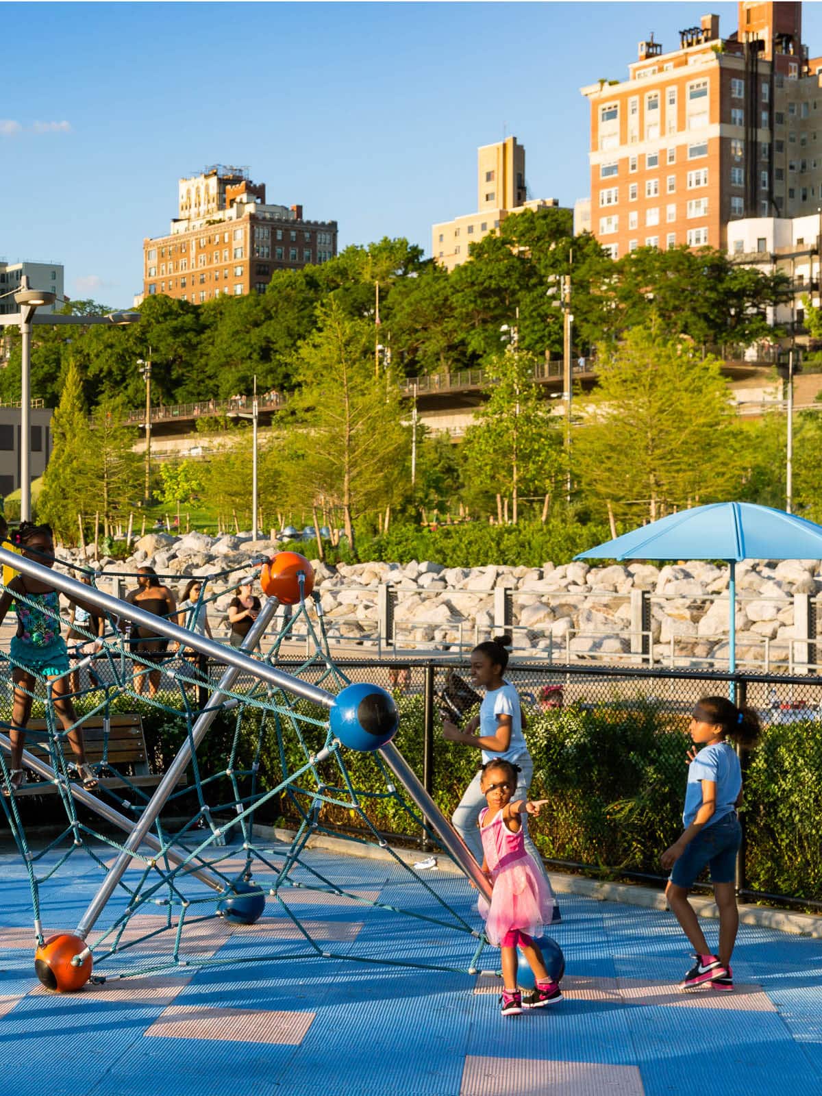 Children playing on climbing structure in a playground at sunset. Brooklyn Heights is seen in the background.