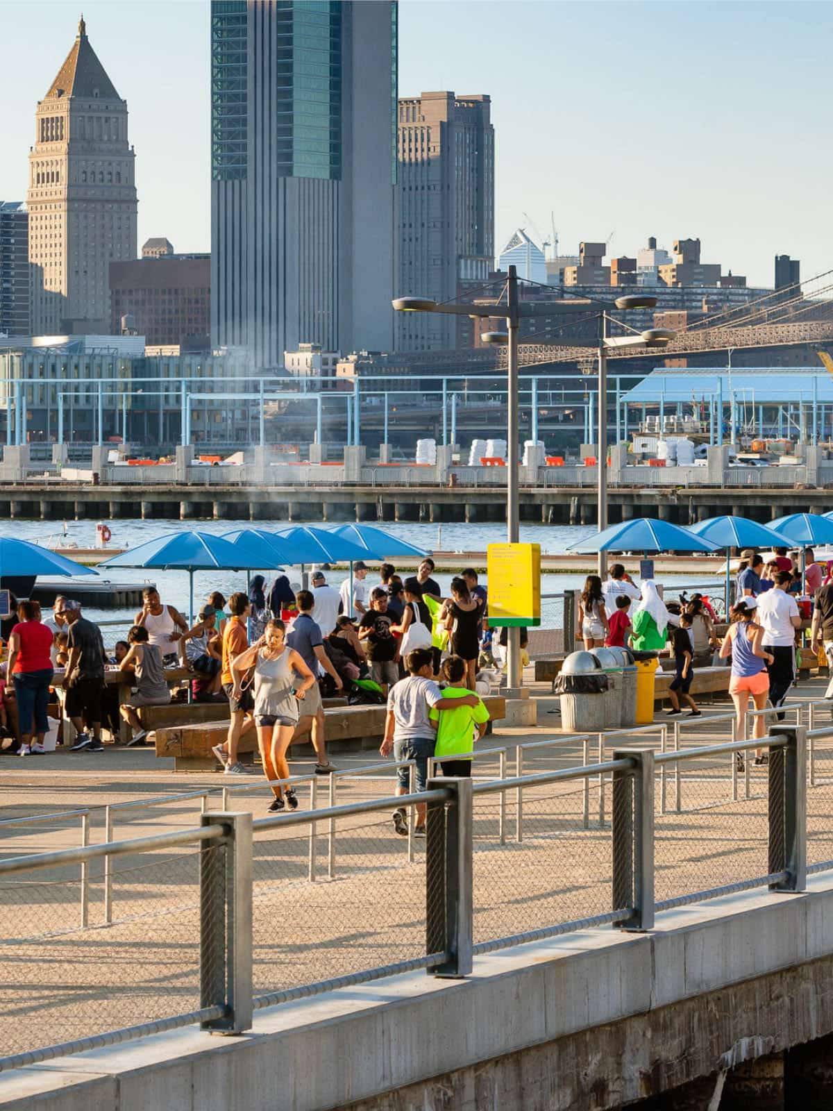People walking by picnic tables at sunset with lower Manhattan in the background.