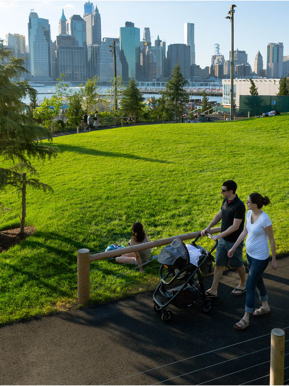 Couple walking with stroller down path beside a lawn at sunset. Lower Manhattan is seen in the background.