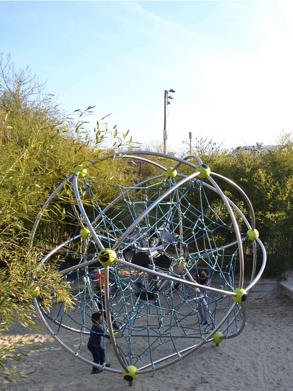 Children playing on round mental climbing structure surrounded by bushes at sunset.