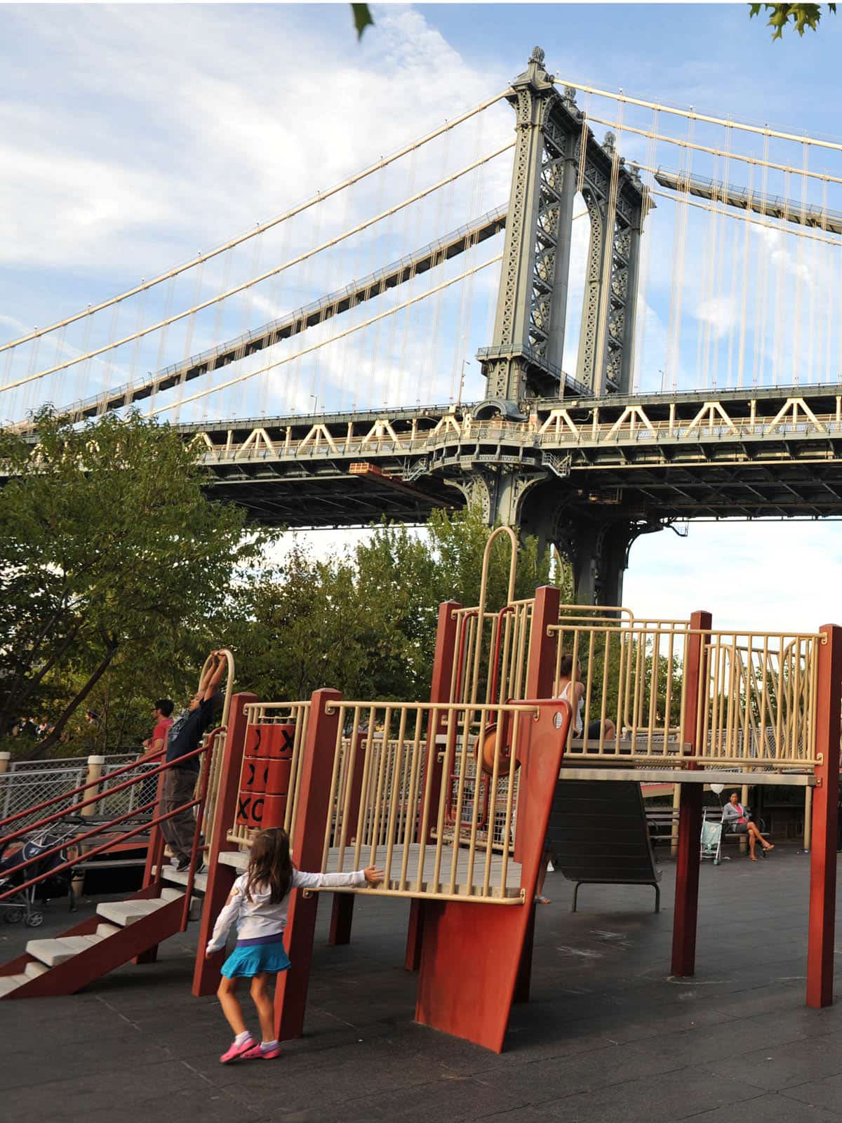 Children playing on a playground structure on a cloudy day. The Manhattan Bridge is seen overhead.