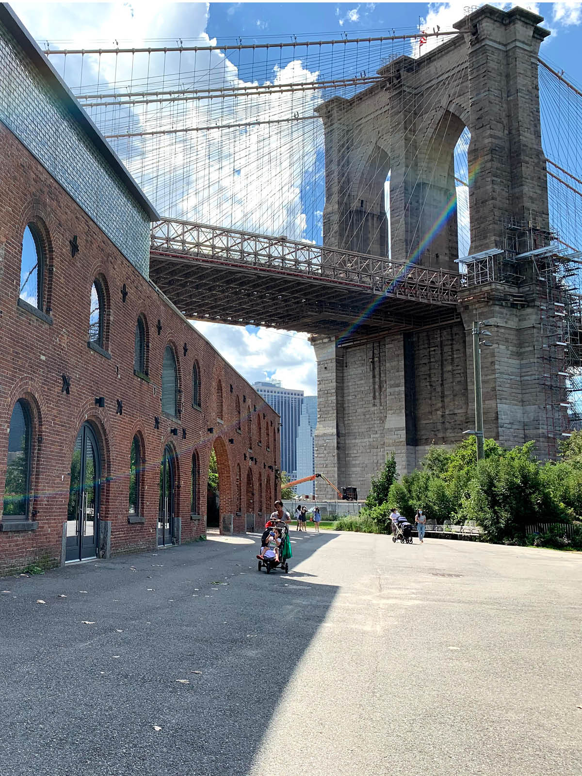 People walking by St Ann's Warehouse on a sunny day. The Brooklyn Bridge is seen overhead.
