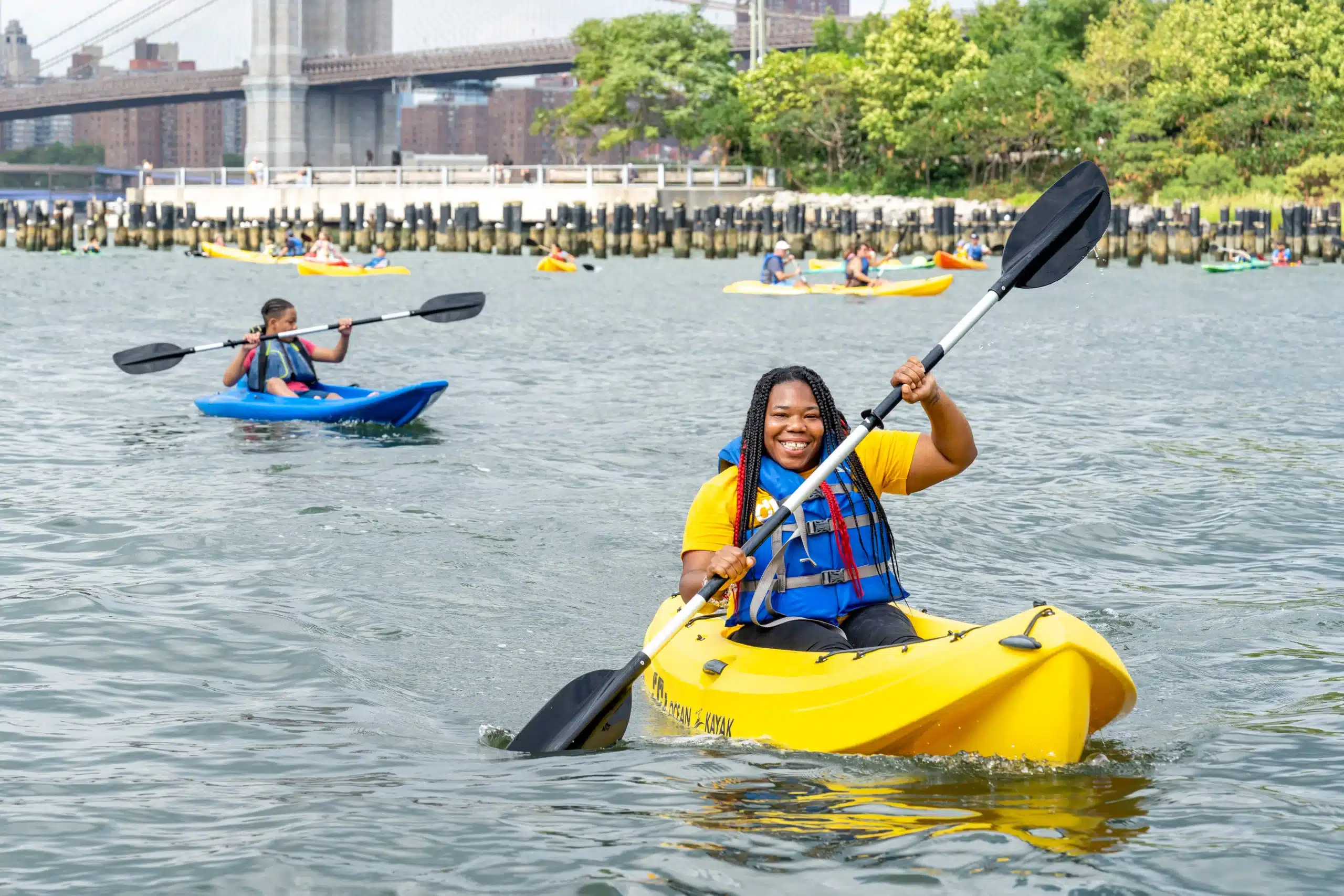 Women kayaking and smiling at camera with piles and Brooklyn Bridge behind